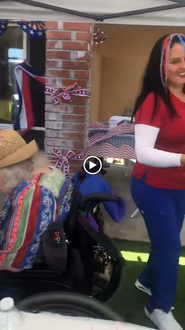 An elderly person in a wheelchair wearing a colorful striped jacket and a straw hat, sitting under a canopy decorated with patriotic ribbons and flags. A woman in a red shirt and blue pants stands nearby, smiling. The setting appears to be an outdoor event or celebration with a brick wall and window in the background.