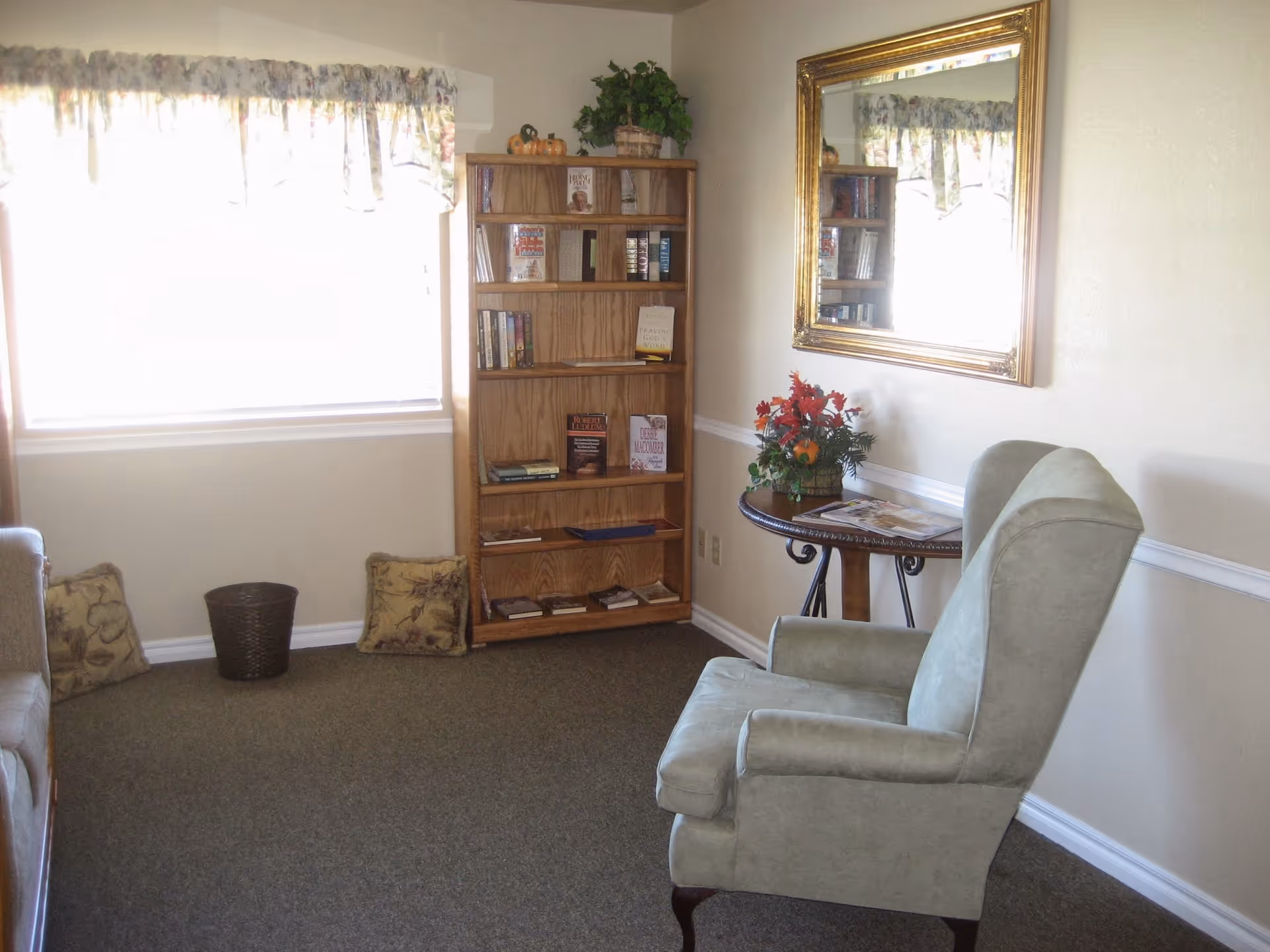 A cozy sitting area with a light green upholstered armchair facing a small round wooden table with a floral arrangement on top. Behind the table is a large gold-framed mirror on the wall. To the left, there is a wooden bookshelf filled with books and decorative items. A window with floral curtains lets in bright natural light, and there are two decorative pillows and a small basket on the floor near the window.