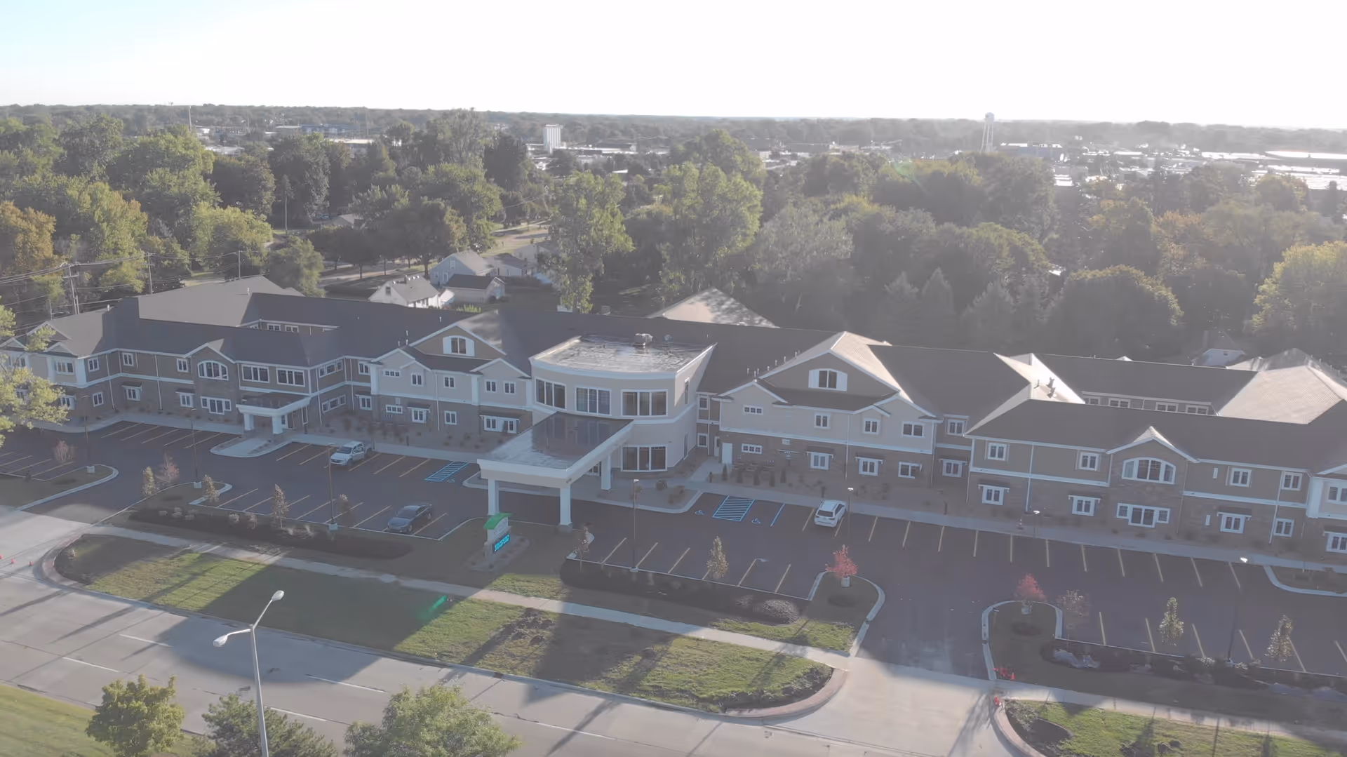 Aerial view of a large senior living facility building with multiple windows and a covered entrance. The building is surrounded by parking spaces, trees, and greenery, with a road in the foreground and a suburban area in the background.