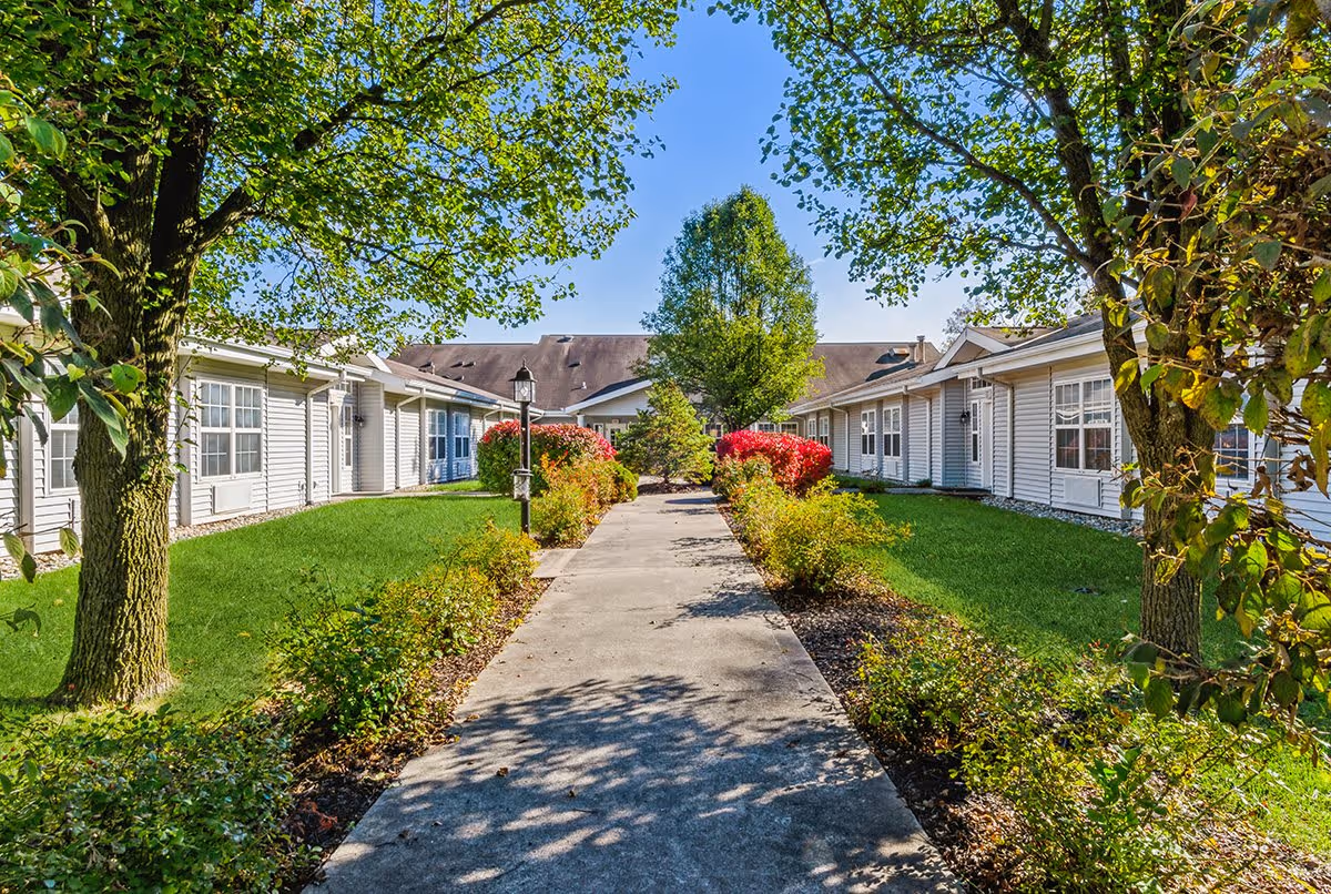 A sunny outdoor courtyard at Cedar Creek of Fort Wayne featuring a concrete walkway lined with green grass, bushes, and trees. Single-story white buildings with multiple windows surround the courtyard under a clear blue sky.