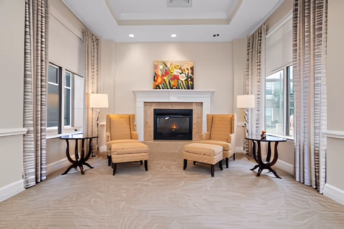 Bright common living area with two armchairs and ottomans facing a fireplace flanked by side tables and lamps.