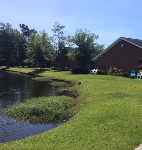 A peaceful outdoor scene at Garden Manor Village featuring a grassy lawn beside a calm pond. There are several trees and shrubs along the water's edge, and brick buildings with white chairs placed outside. The sky is clear and blue.