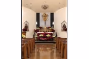 Interior view of a chapel with wooden pews on both sides leading to an altar decorated with flowers and a large crucifix mounted on the wall behind it.
