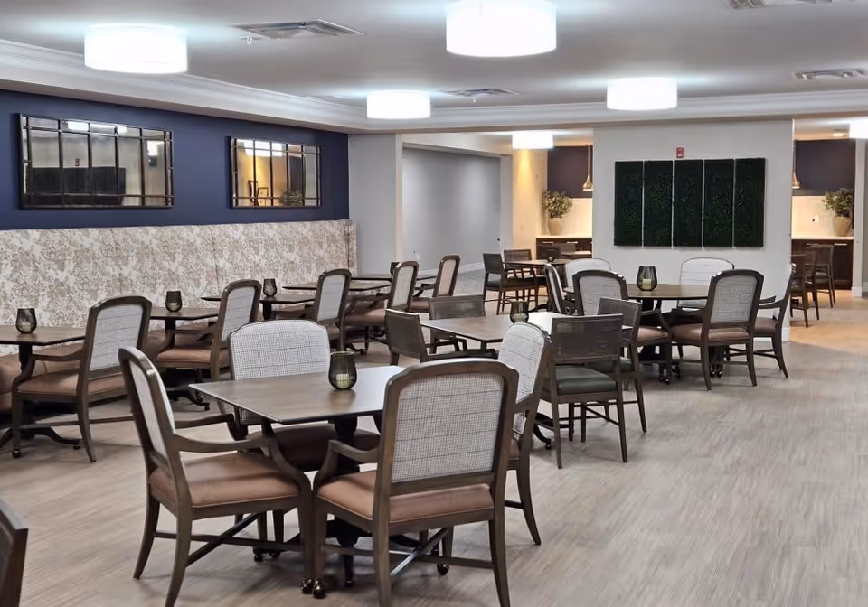 Well-lit communal dining room with multiple tables and chairs arranged on wood-look floors and modern ceiling lights.