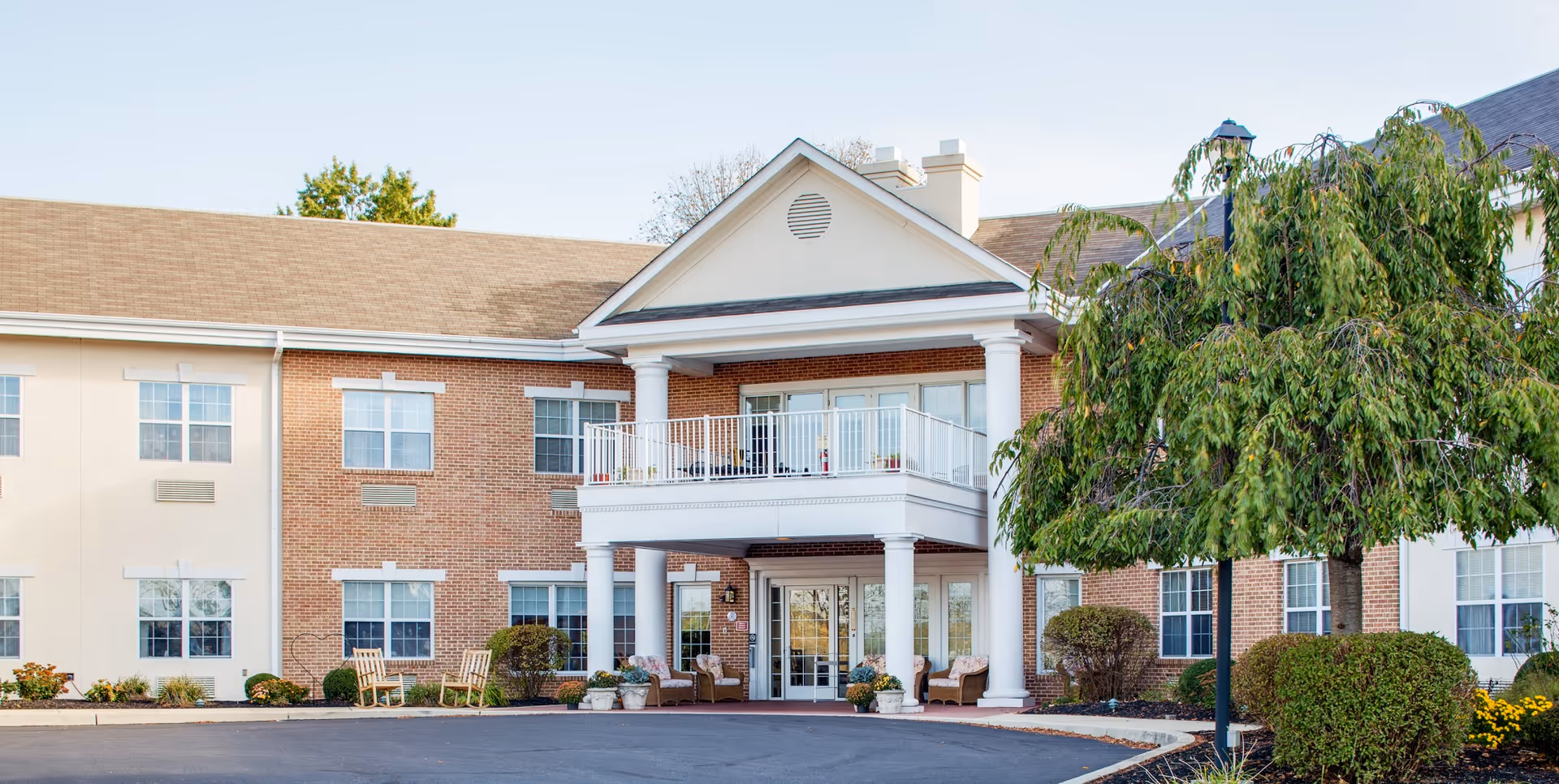 Front exterior view of a two-story senior living facility building with a covered entrance supported by white columns, a balcony above the entrance, multiple windows, and landscaped bushes and trees around the building.