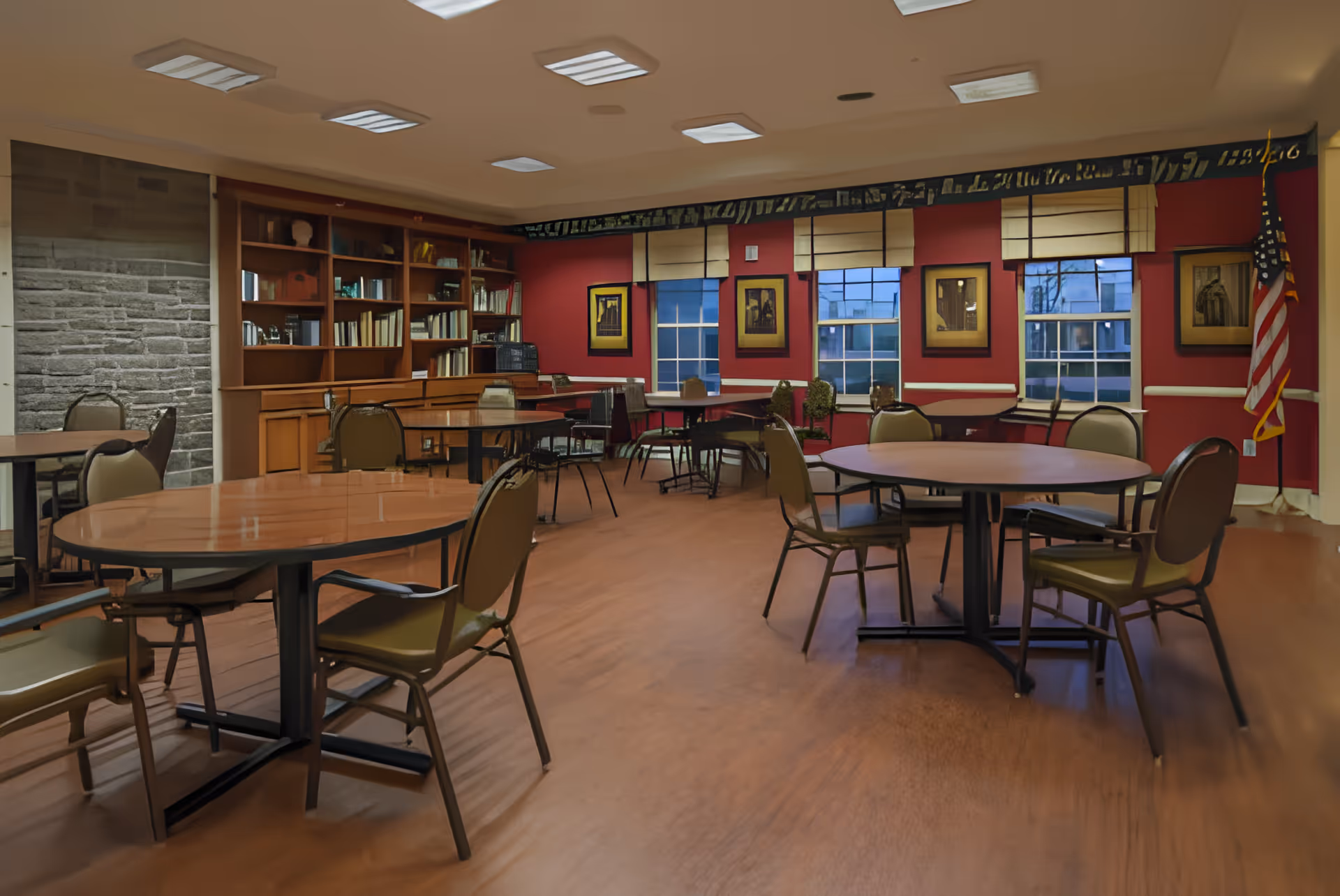 A spacious room with multiple round and rectangular tables surrounded by chairs. The walls are painted red and decorated with framed pictures. There are several windows with beige blinds, a bookshelf filled with books, and an American flag in the corner. The floor is wooden, and the ceiling has recessed lighting.