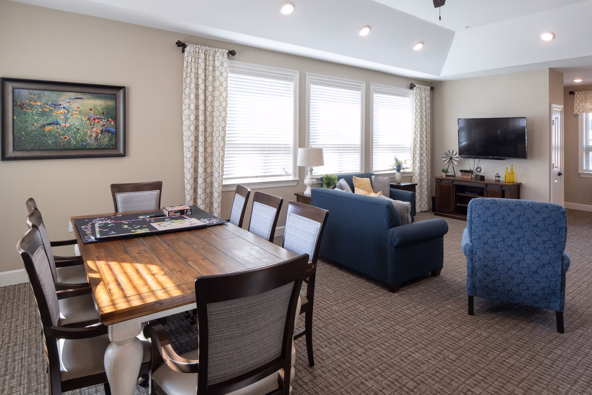 A bright and spacious living and dining area in an assisted living facility. The dining area features a wooden table with six chairs, a puzzle in progress on the table, and a framed picture of flowers on the wall. The living area has a blue sofa, a blue armchair, a TV mounted on the wall, and a wooden TV stand with decorative items. Large windows with patterned curtains allow natural light to fill the room.