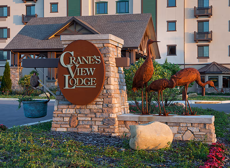 Stone sign for Cranes View Lodge with metal sculptures of cranes beside it, set in landscaped greenery with a multi-story building in the background.