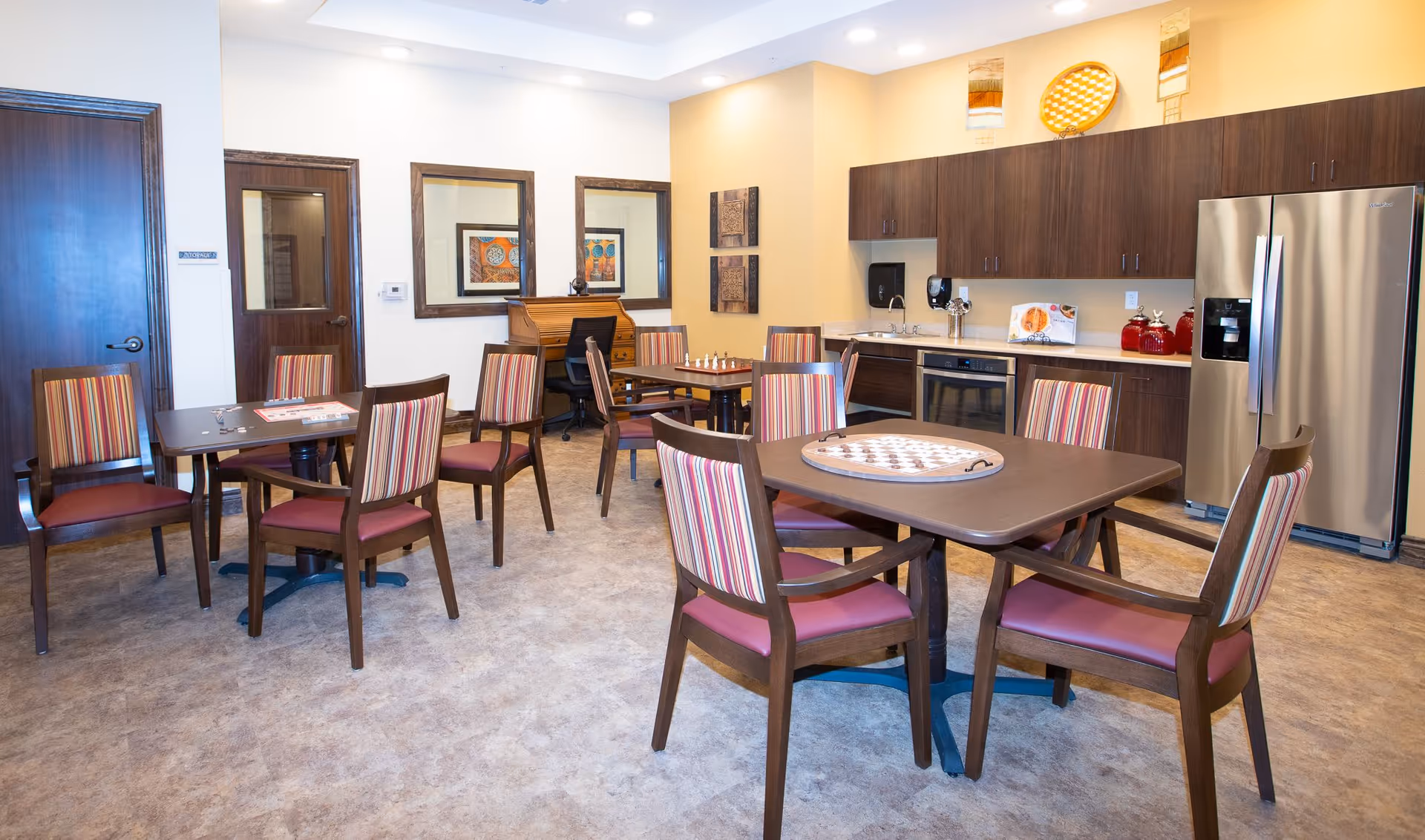 Bright communal dining/activity room with several tables and striped-upholstery chairs, a kitchenette with a stainless-steel refrigerator, and board games on the tables.