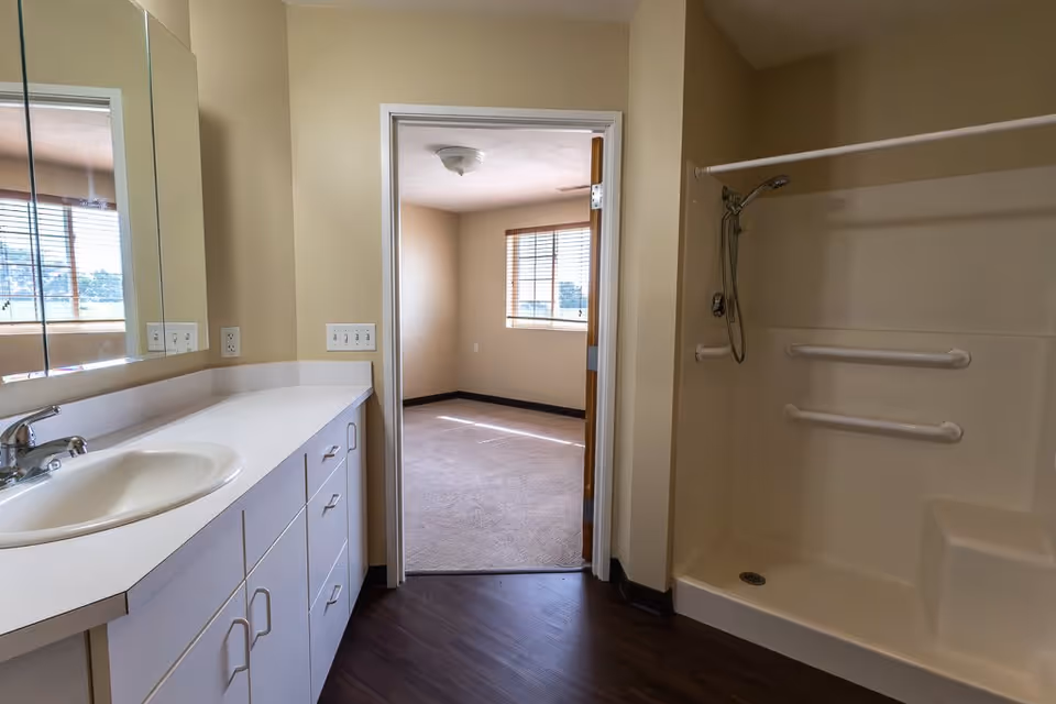 Interior view of a bathroom in a senior living facility showing a white sink with a countertop and cabinets underneath on the left, a large mirror above the sink, and a walk-in shower with grab bars and a handheld showerhead on the right. Through an open door, a carpeted bedroom with a window and blinds is visible.