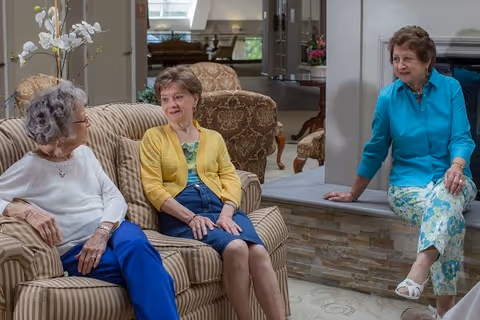 Three elderly women sitting and chatting in a cozy living room area with comfortable chairs and a fireplace in the background.