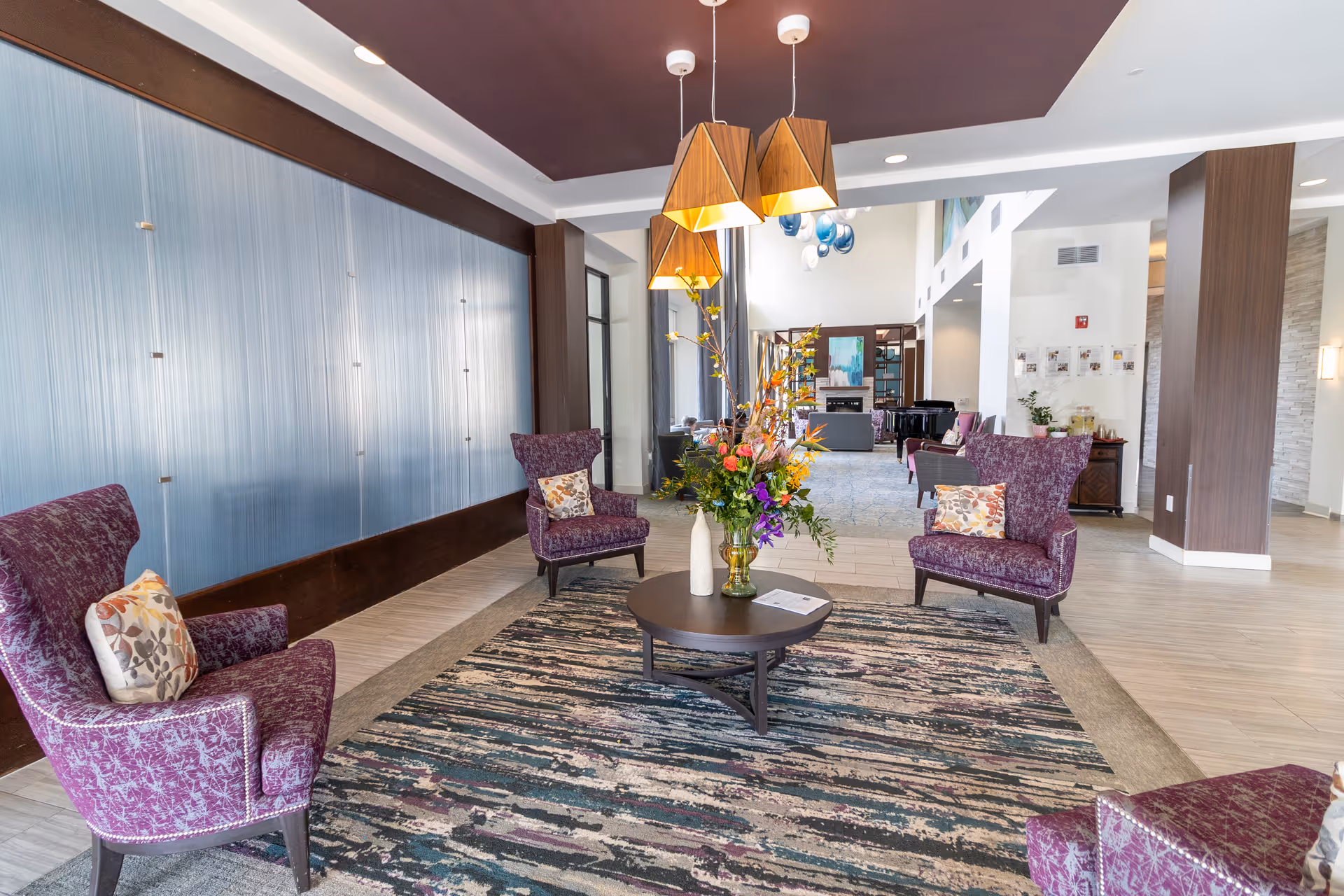 Lobby seating area with purple armchairs arranged around a round coffee table topped with a flower arrangement under pendant lights.