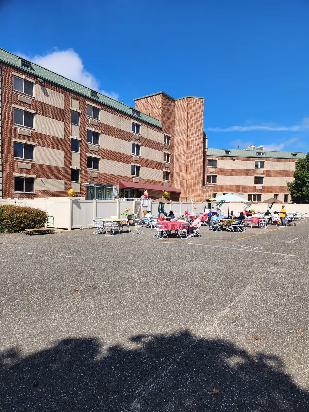 Outdoor area of a senior living facility with several tables and chairs set up on a paved surface. People are seated at the tables under umbrellas, enjoying the sunny weather. The background shows a multi-story brick building with a green roof under a clear blue sky.