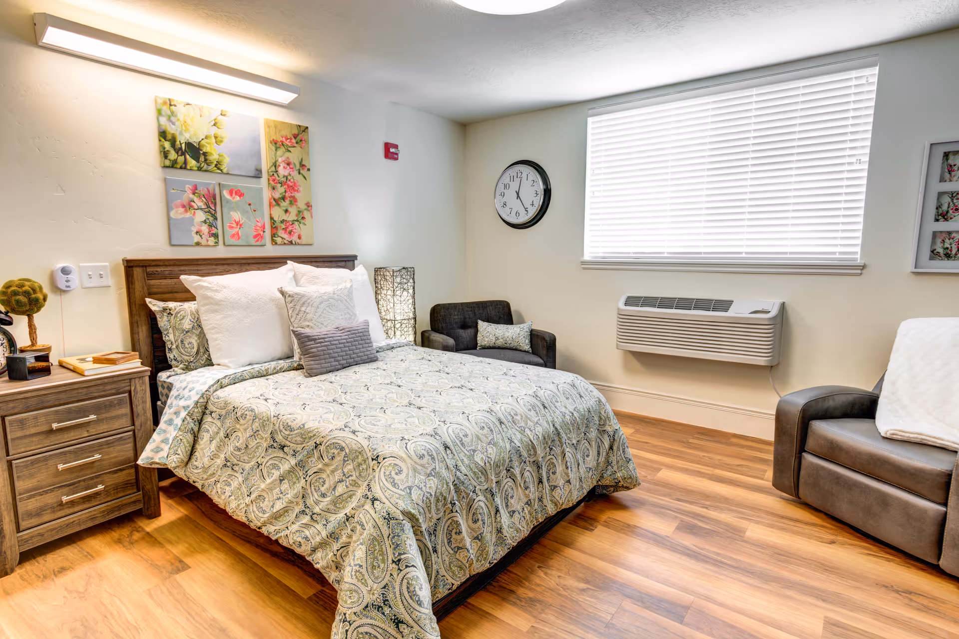 A cozy bedroom with a wooden bed frame, patterned bedspread, and multiple pillows. There is a wooden nightstand with books and a decorative item, a wall clock, two armchairs, a window with blinds, and floral artwork on the wall.