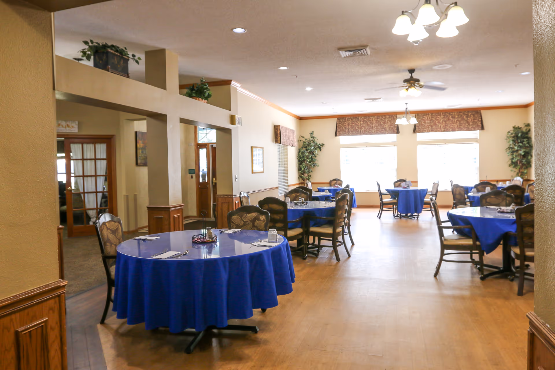 Dining room with multiple round tables covered with blue tablecloths, each surrounded by chairs. The room has large windows with valances, ceiling fans, and light fixtures. There are plants placed near the windows and decorative elements on the walls.