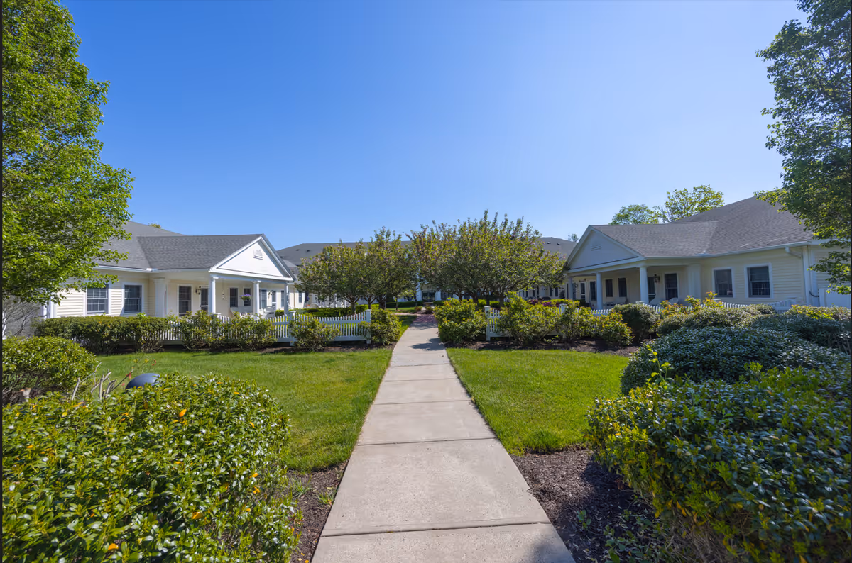 A paved walkway flanked by green bushes and trees leads through a well-maintained garden area between two single-story white buildings with porches under a clear blue sky.