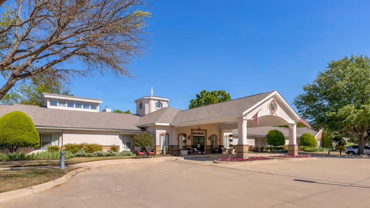Exterior view of Brookdale Stonebridge Ranch facility showing a single-story building with a covered entrance, manicured landscaping, and clear blue sky.