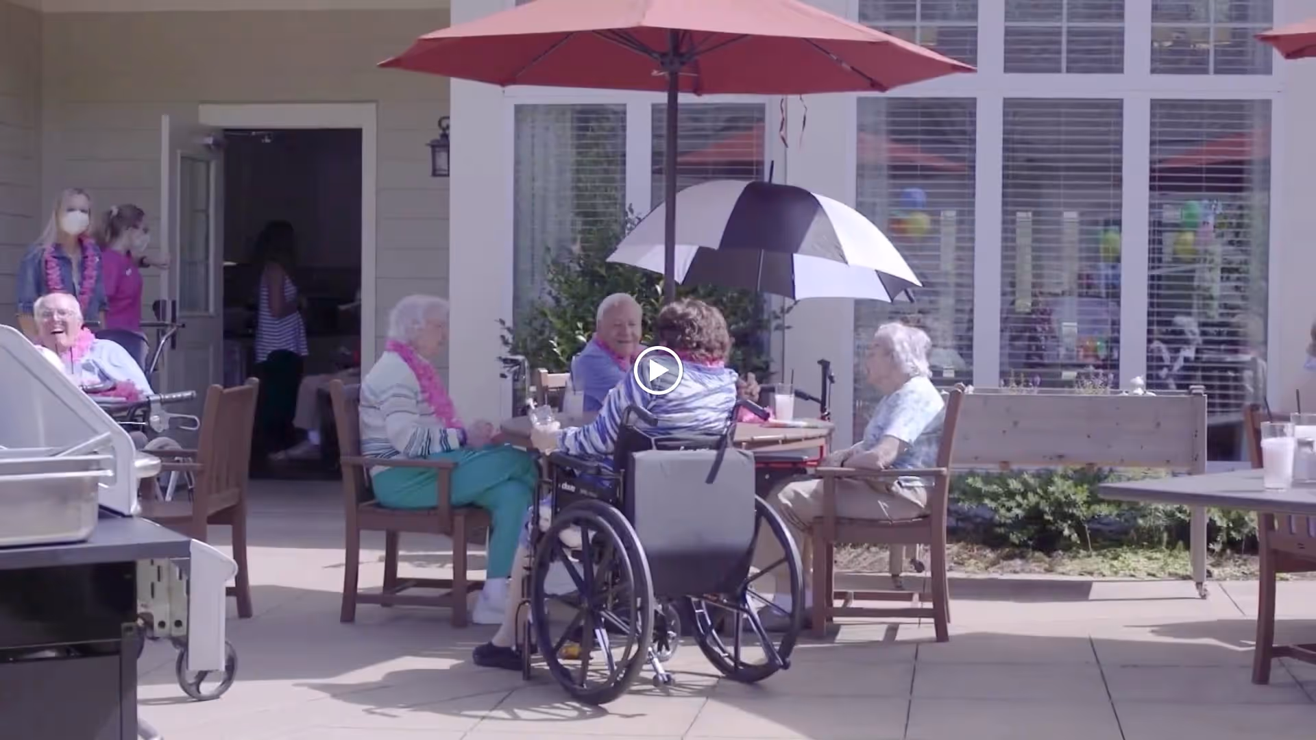 A group of seniors seated around a patio table under umbrellas outside a senior living facility, with a wheelchair visible.