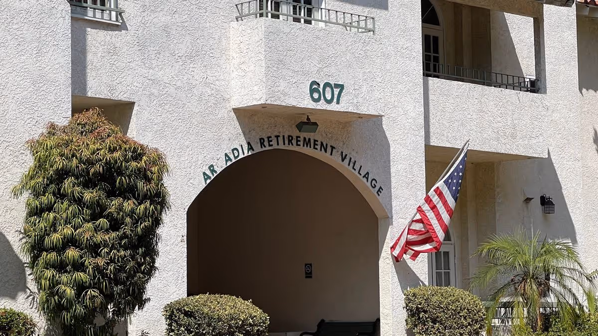Entrance arch and facade of Arcadia Retirement Village showing address number 607, an American flag, and surrounding shrubs.