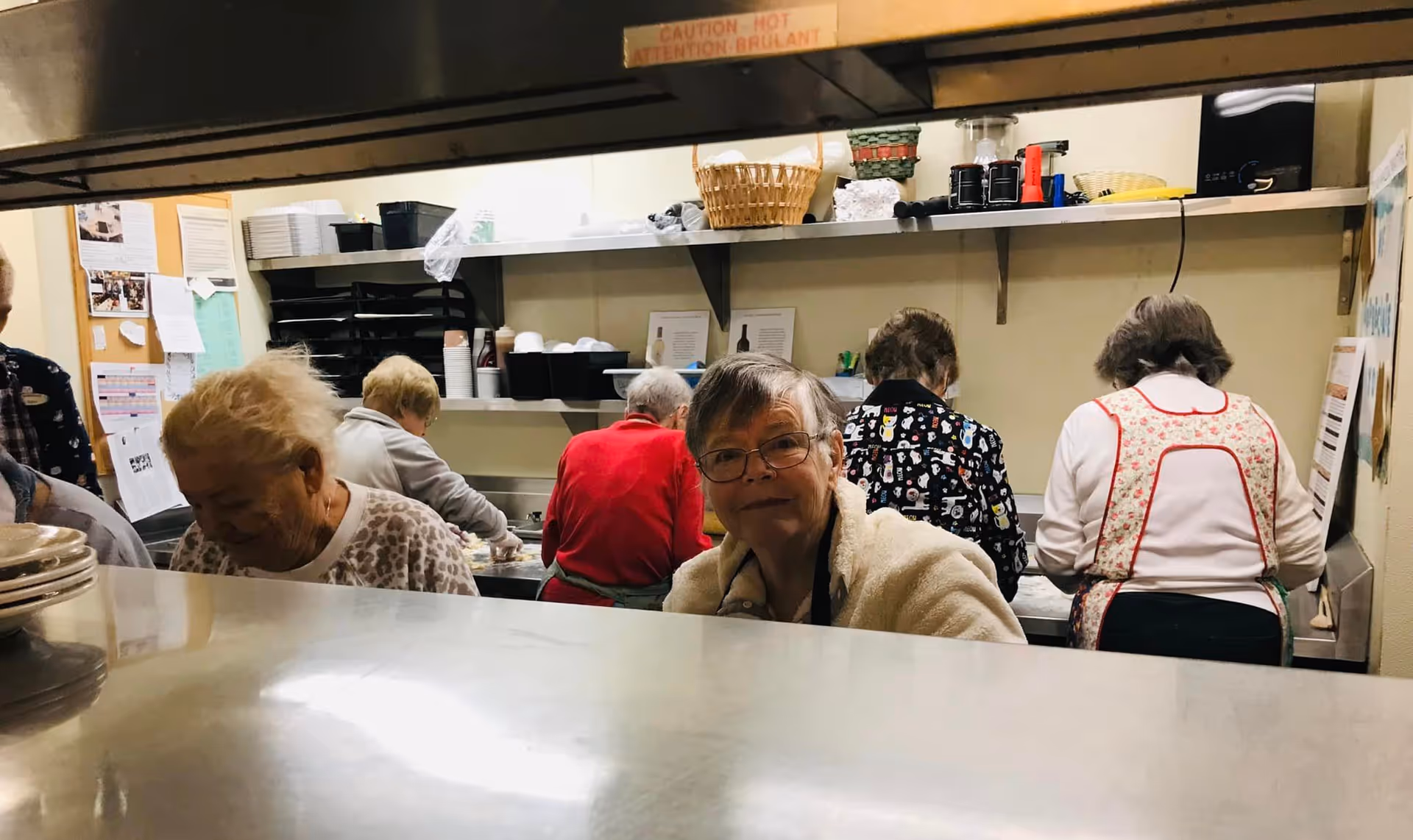A group of elderly women working together in a kitchen area, some wearing aprons, with shelves holding various kitchen items and a bulletin board in the background.
