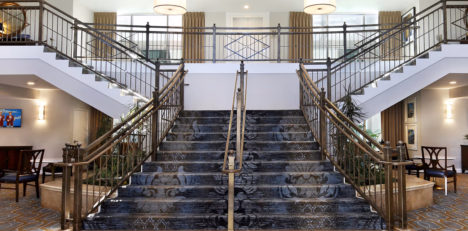 Grand lobby interior with a wide carpeted double staircase, metal railings, seating areas and large windows.