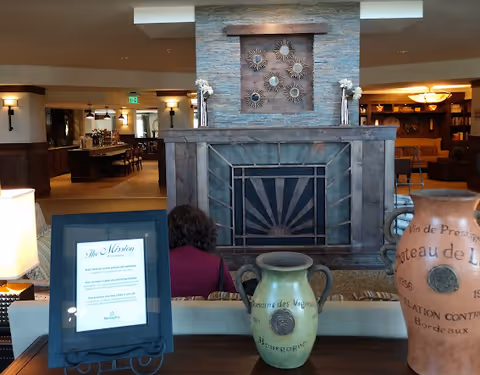 Interior view of a cozy assisted living facility common area featuring a large stone fireplace with decorative sunburst metal art above it. In front of the fireplace, there is a wooden table with two large decorative vases and a framed mission statement. A person with curly hair is seated facing the fireplace. The background shows a well-lit dining area with tables and chairs.