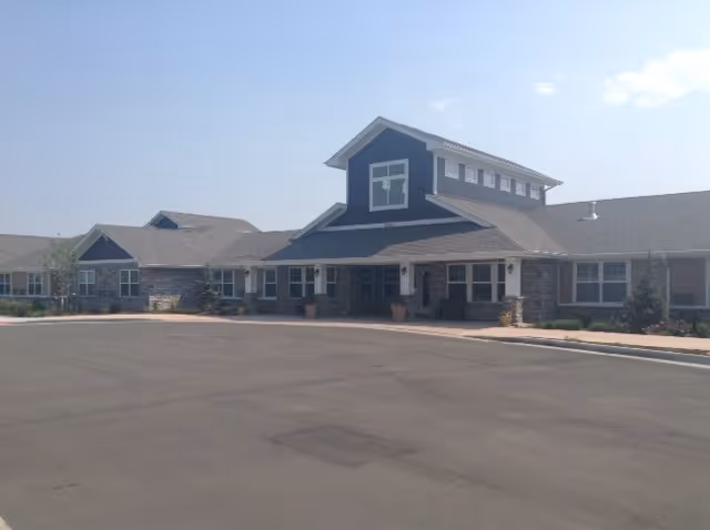 Exterior view of a single-story building with a large paved driveway in front. The building has a combination of stone and siding on the facade, multiple windows, and a prominent central section with a gabled roof and additional upper windows. The sky is clear with a few clouds.
