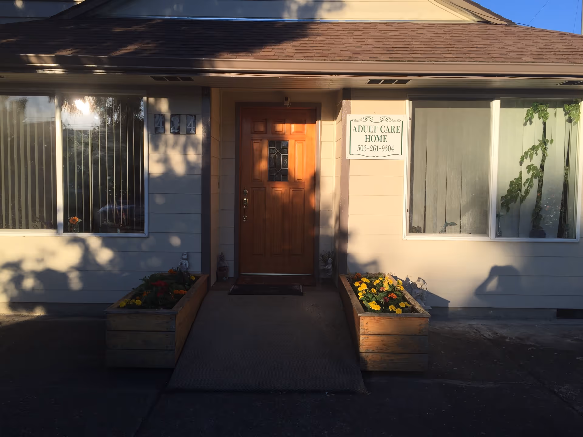Front entrance of a single-story adult care home with a wooden door, two large windows, flower planters, and a sign reading 'Adult Care Home'.