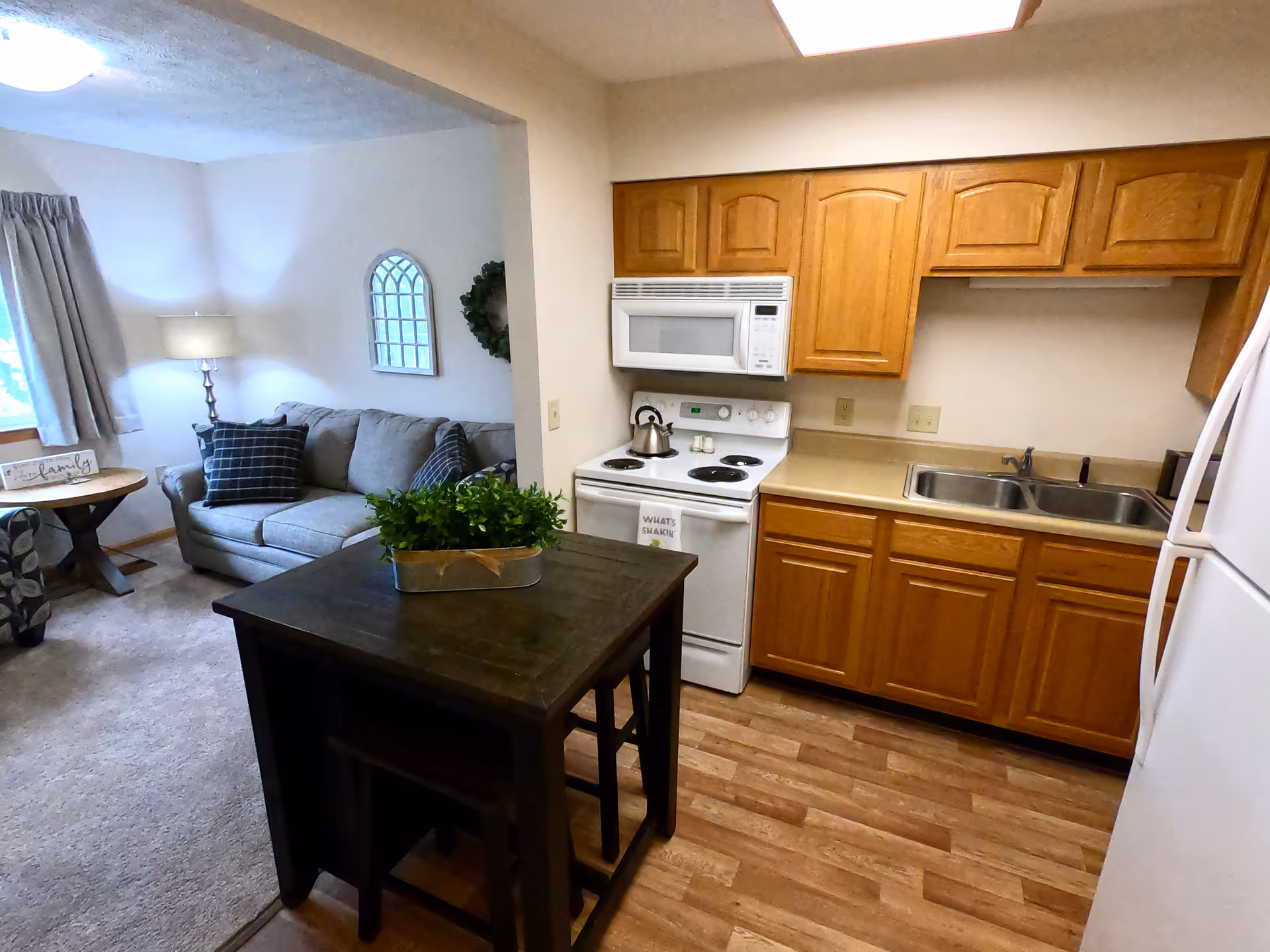 A cozy living space featuring a small kitchen with wooden cabinets, a white stove, microwave, double sink, and refrigerator. Adjacent to the kitchen is a living room area with a gray sofa, a side table with a lamp, a window with curtains, and decorative wall accents. A dark wooden table with a green plant centerpiece separates the kitchen and living room.