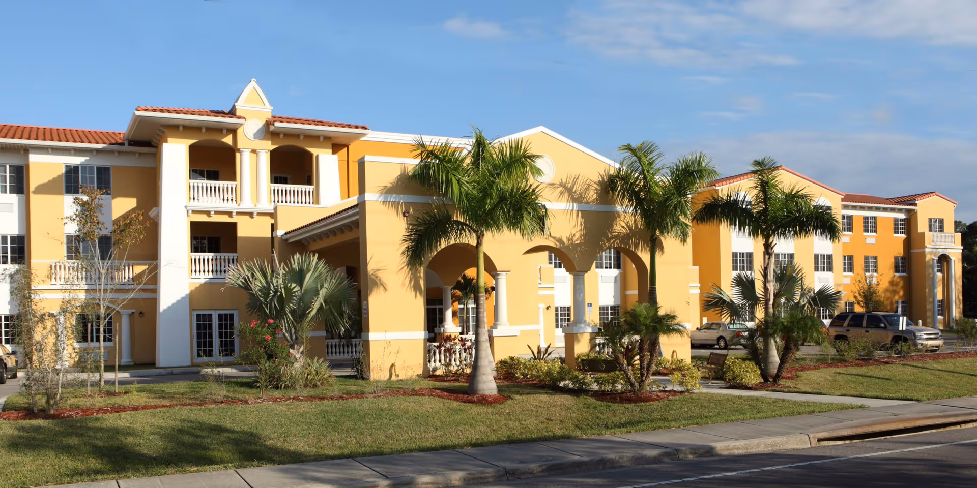 Yellow three-story assisted living building with arched covered entrance, balconies, palm trees and landscaped lawn.