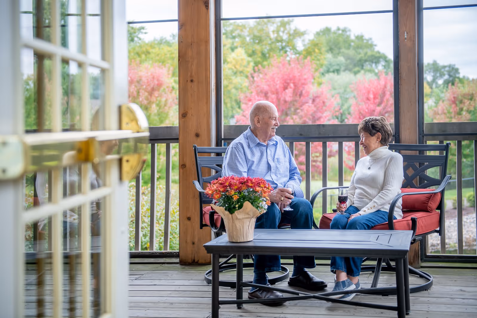 An elderly man and woman sitting on a covered outdoor porch or patio, engaged in conversation and holding glasses of red wine. The porch has wooden flooring and railing, with colorful autumn trees visible in the background. A table with a potted plant featuring red and orange flowers is in front of them.