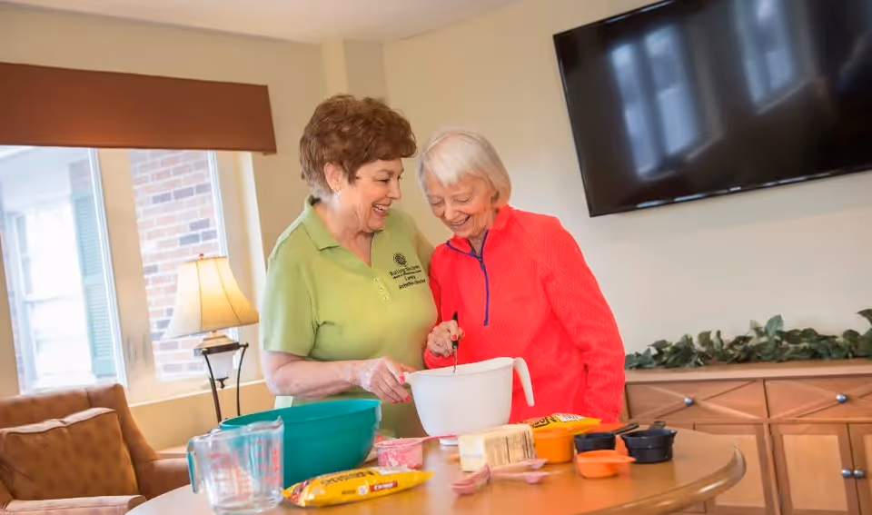 Two elderly women smiling and mixing ingredients in a white bowl on a table in a cozy living room setting. The table has various baking ingredients and measuring cups. A large flat-screen TV is mounted on the wall behind them, and a window with a lamp and a brown couch are visible in the background.