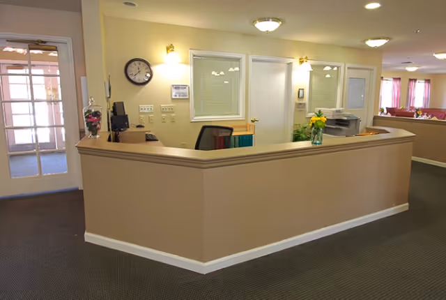 A beige reception desk in the lobby of a senior living facility with chairs, a wall clock, and vases of flowers.