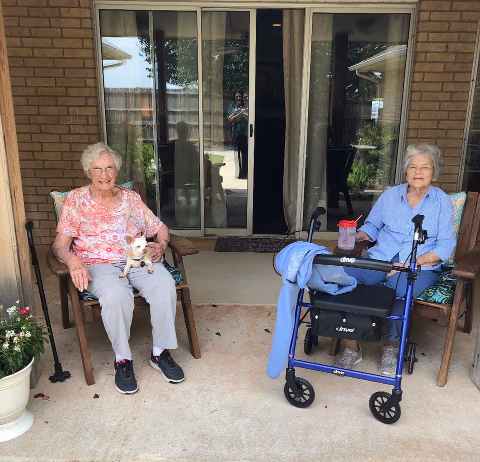 Two elderly women sitting on wooden chairs on a covered patio outside a brick building. One woman is holding a small white dog on her lap, and the other has a blue walker with a jacket draped over it and a cup with a straw on the armrest. There is a potted plant on the left side and a sliding glass door behind them reflecting a person taking the photo.