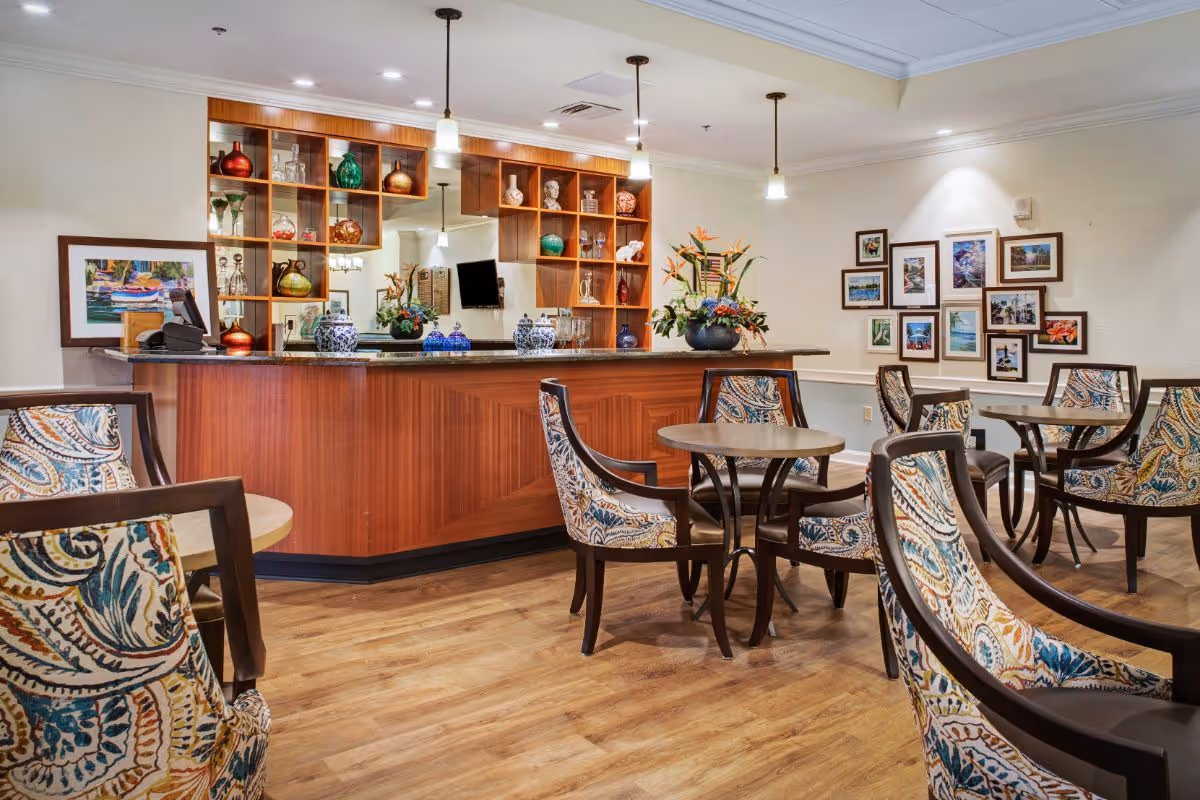 A cozy common area with a wooden bar counter decorated with vases and glassware. Several round tables with patterned upholstered chairs are arranged on a wooden floor. The walls feature framed pictures and the ceiling has recessed lighting and hanging pendant lights.