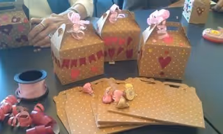 A table with several small brown gift boxes decorated with pink ribbons and heart designs, along with matching brown paper bags and pink curling ribbon. A person's hands are visible arranging the boxes.