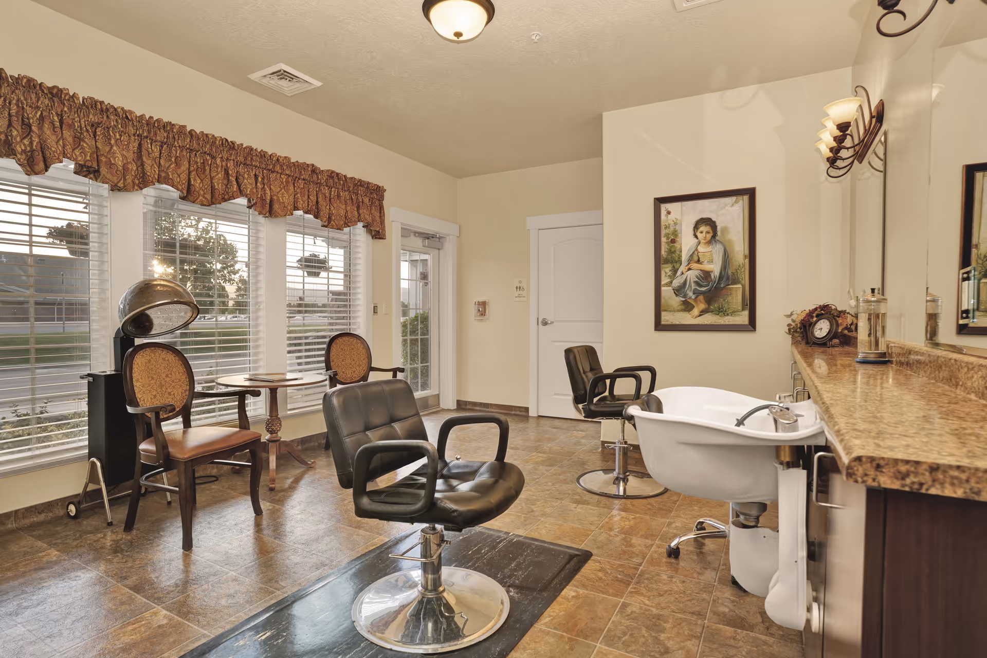 Interior of a salon area with two black salon chairs, a white hair washing sink, two wooden chairs with patterned upholstery, a small round table, large windows with blinds and valances, a painting of a girl on the wall, and a countertop with various items.