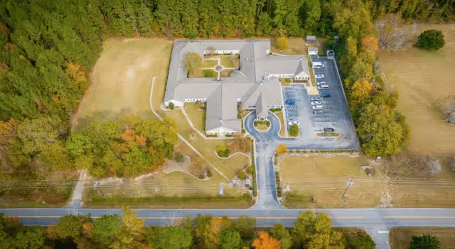 Aerial view of The Legacy of Orangeburg senior living facility showing a large building with a circular driveway, parking lot with several cars, surrounding trees, and open grassy areas adjacent to a road.