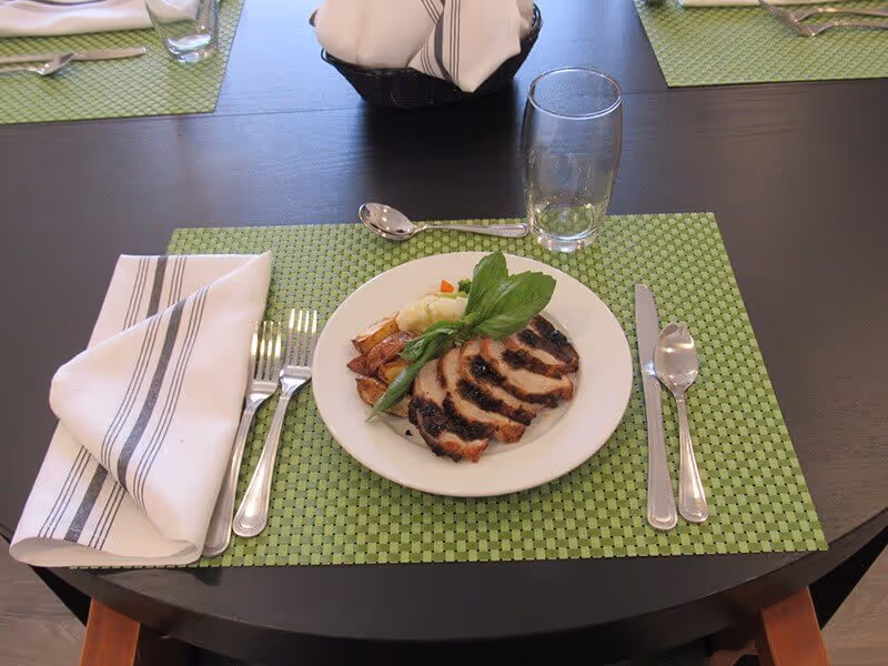 A plated serving of sliced roasted meat and vegetables on a green placemat at a set dining table with napkin, utensils, and a glass.