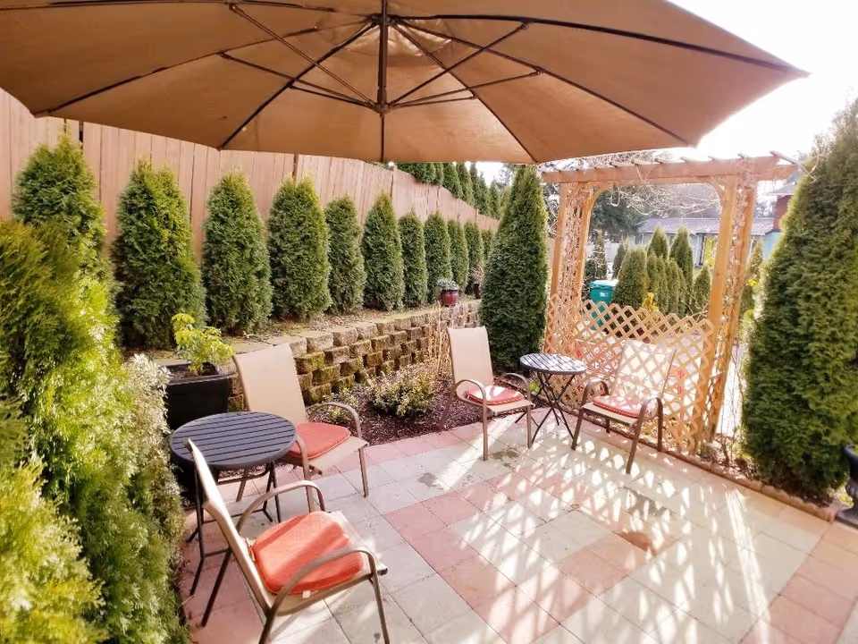 Outdoor patio area with a large brown umbrella, several chairs with red cushions, two small round tables, surrounded by tall green shrubs and a wooden fence. There is a wooden trellis at the far end of the patio.