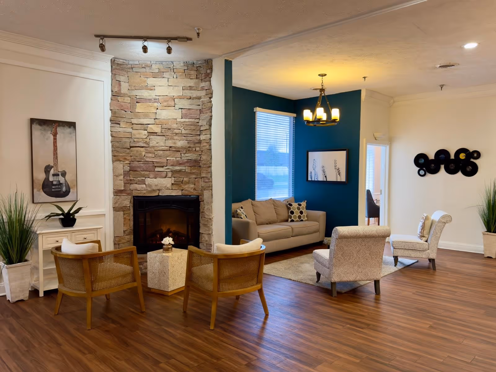 A cozy living room area with a stone fireplace in the center. Two wooden chairs with cushions face the fireplace, and a small square table is placed between them. To the right, there is a beige sofa with patterned pillows against a teal accent wall, under a chandelier light fixture. Two patterned armchairs are positioned on a rug in front of the sofa. The room has wooden flooring, potted plants, and wall decorations including a guitar painting and a vinyl record display.