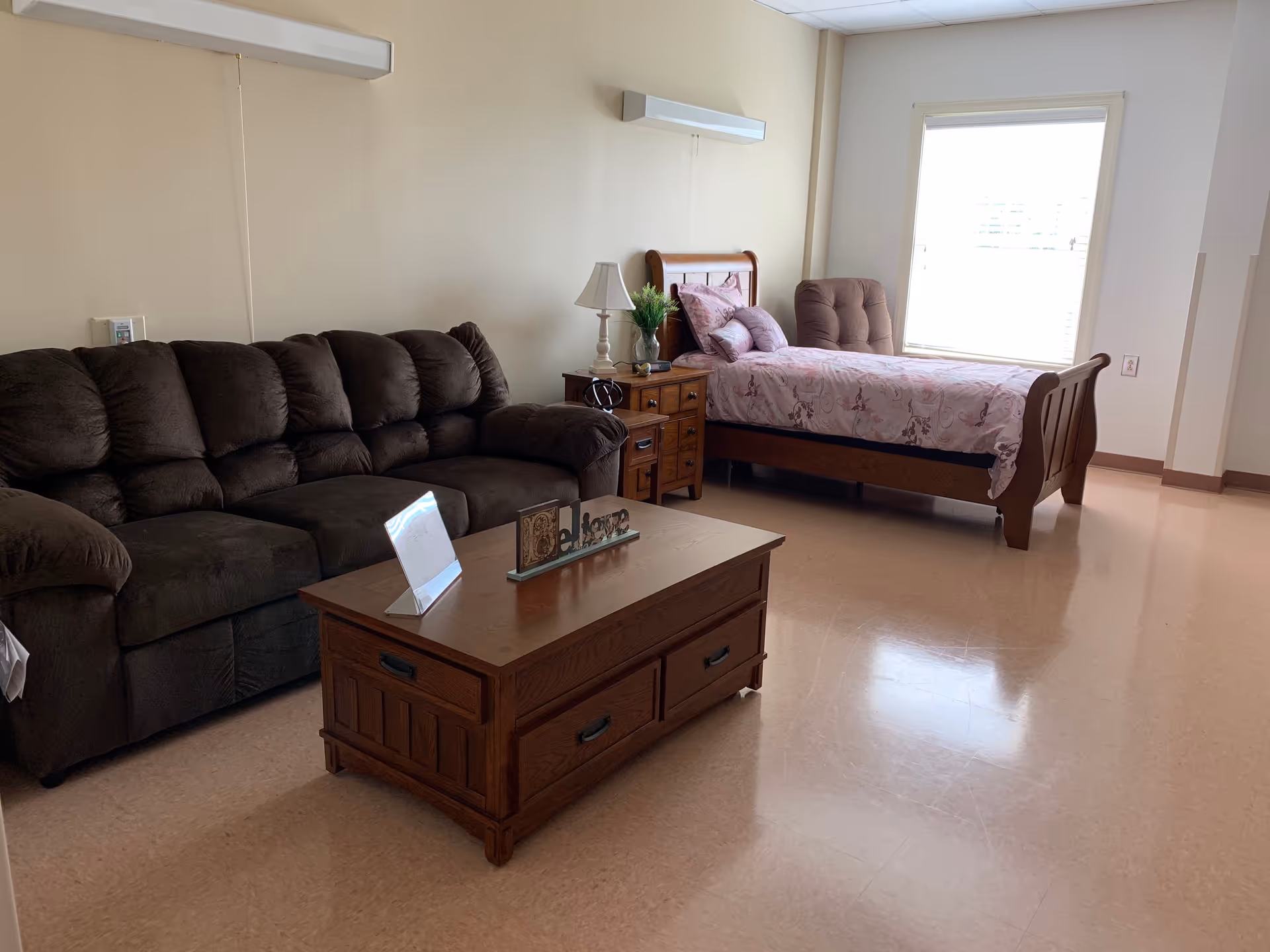 A room with a dark brown plush sofa, a wooden coffee table with a decorative welcome sign, a wooden nightstand with a lamp and a plant, and a single bed with floral bedding and a cushioned chair near a window.