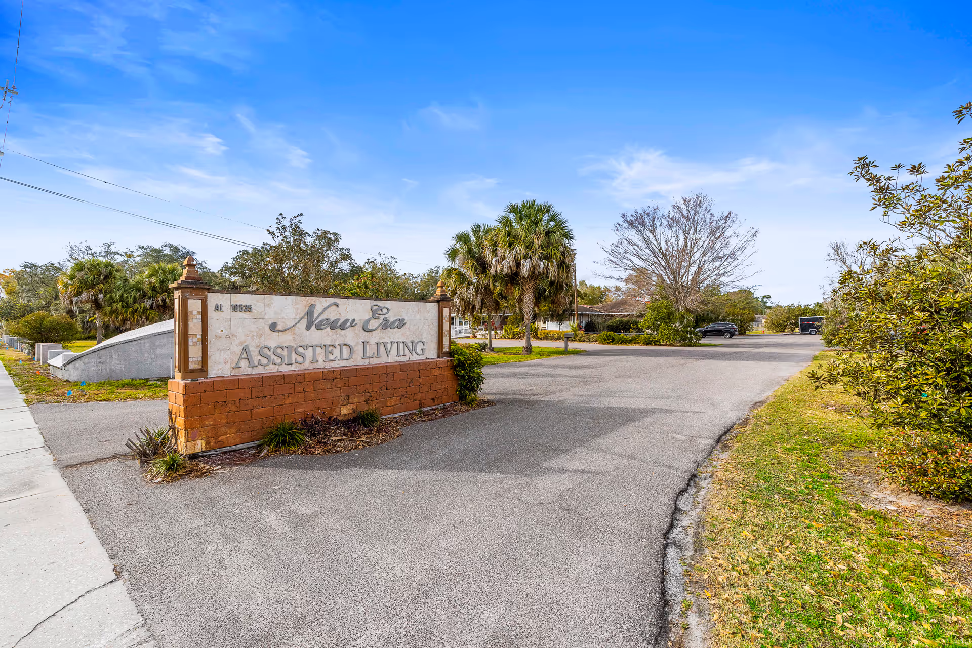 Entrance sign for New Era Assisted Living made of brick and stone, situated next to a paved driveway with trees and bushes on either side under a blue sky.