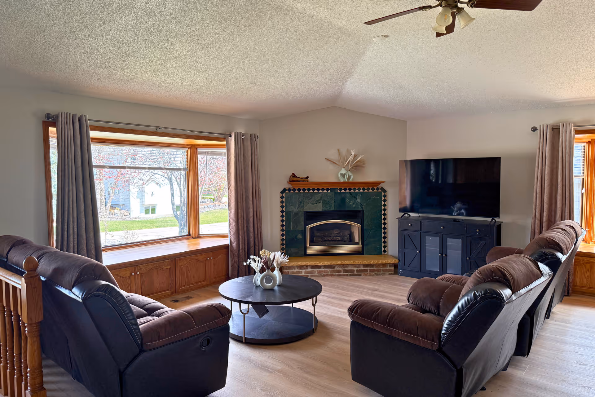 A cozy living room with two brown reclining sofas facing a round coffee table with a decorative vase. The room features a corner fireplace with green and black tile surround, a large flat-screen TV on a black cabinet, and two large windows with gray curtains letting in natural light. The floor is light wood, and a ceiling fan with lights is visible above.