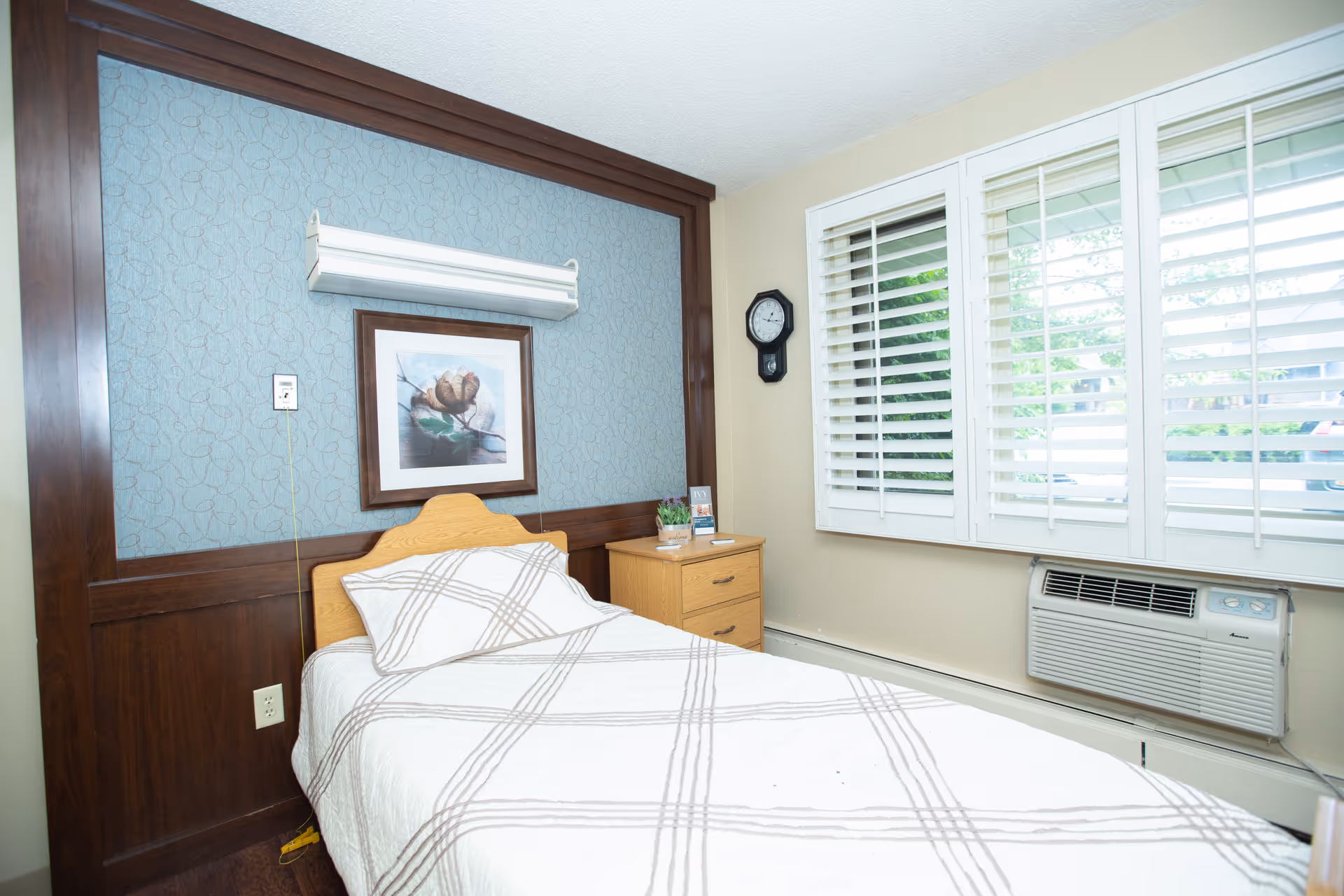 A small bedroom with a single bed covered in a white and beige plaid bedspread. The bed has a wooden headboard and is positioned against a wall with blue patterned wallpaper and dark wood paneling. Above the bed is a framed picture of a flower and a wall-mounted light fixture. To the right of the bed is a wooden nightstand with a small plant and some items on top. A wall clock is mounted near a window with white plantation shutters, and below the window is a wall-mounted air conditioning unit.