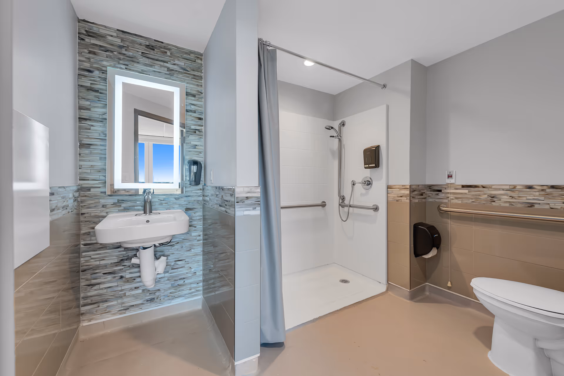 Accessible bathroom with a wall-mounted sink and illuminated mirror at left, roll-in shower with grab bars in the center, and a toilet at right.
