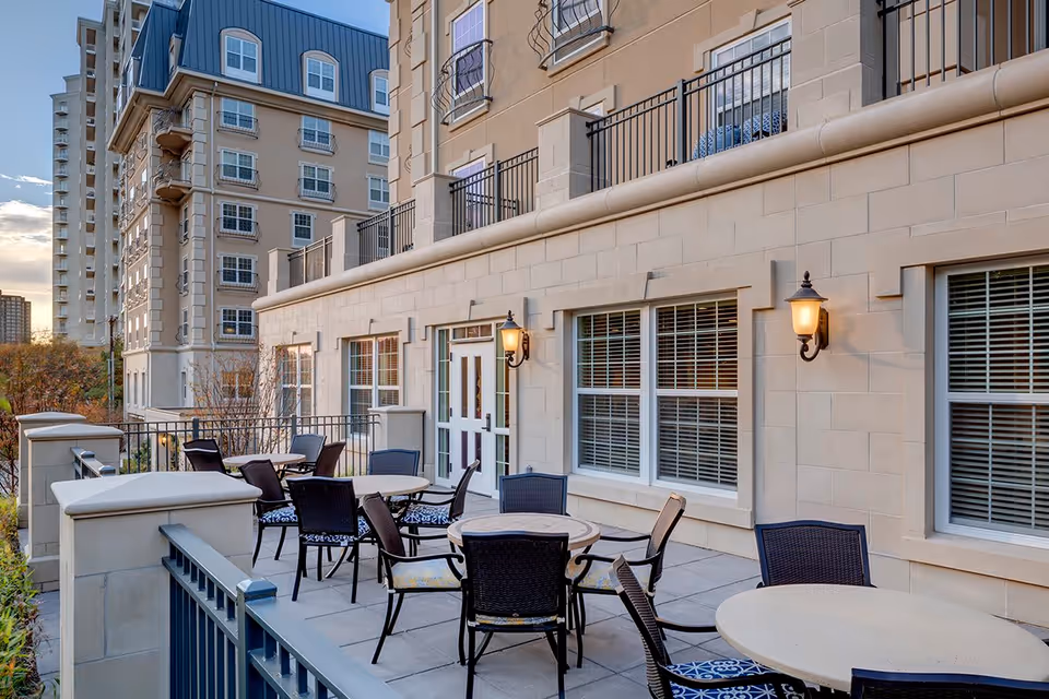 Outdoor patio area at Belmont Village Senior Living Turtle Creek with round tables and chairs arranged for seating. The patio is adjacent to a beige stone building with large windows and wall-mounted lantern-style lights. In the background, there are taller buildings and trees under a partly cloudy sky.