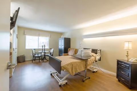 A well-lit senior care facility room with a hospital-style bed, bedside table with a lamp and telephone, a small round table with two chairs near a window, and a tall cabinet. The room has wooden flooring and light-colored walls.