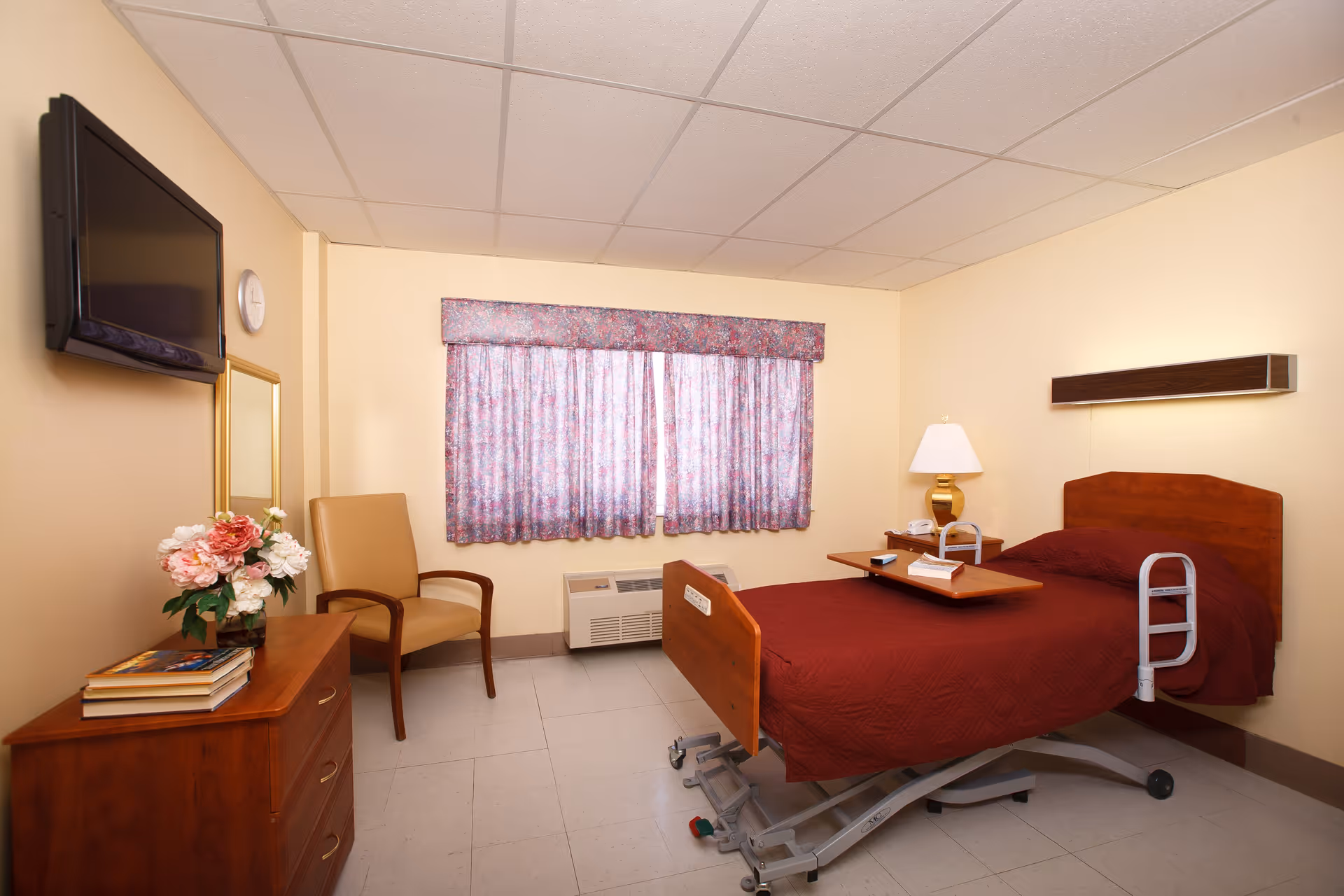 A senior living facility bedroom with a hospital-style adjustable bed covered in a maroon quilt, a wooden bedside table with a lamp and phone, a wooden dresser with books and a vase of flowers, a beige armchair, a wall-mounted TV, a mirror, and a window with floral curtains.