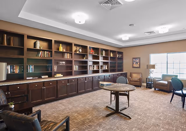 A spacious room with a large built-in wooden bookshelf filled with books and decorative items along one wall. The room has carpeted flooring, a round table with chairs in the center, and several armchairs arranged near a window that lets in natural light. The ceiling has recessed lighting fixtures.